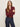 A woman with long light brown hair stands with arms crossed, wearing the Sanctuary Clothing dreamgirl lace button up night berry top and high-waisted blue jeans with a burgundy belt, against a plain light background.