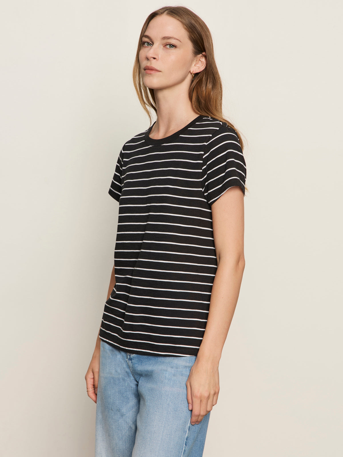 A woman with long, light brown hair wears the Sanctuary Clothing "perfect tee white black stripe 5" and light blue jeans, standing against an off-white background and looking slightly to the side with a neutral expression.