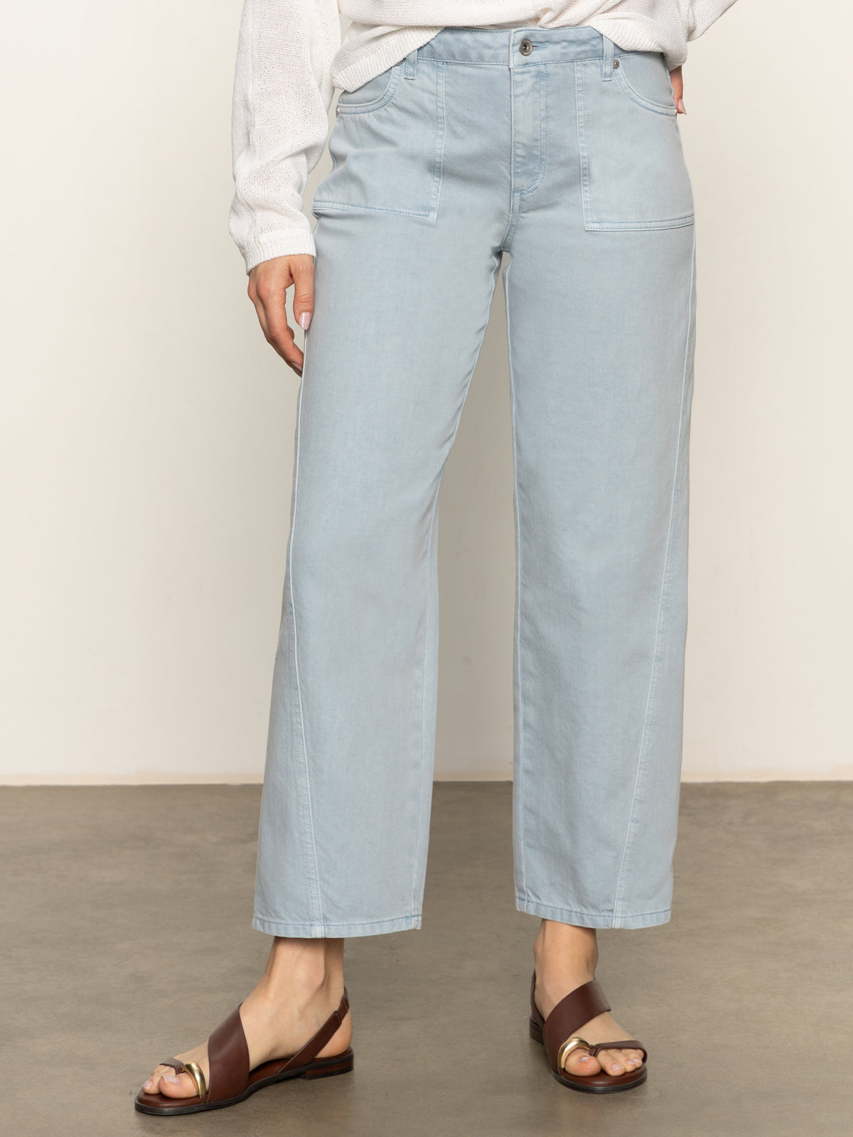 A person wears Sanctuary Clothing’s district column ankle jean in blue mirage with front pockets, a white long-sleeve top, and brown sandals, standing on a gray floor against a plain light background.