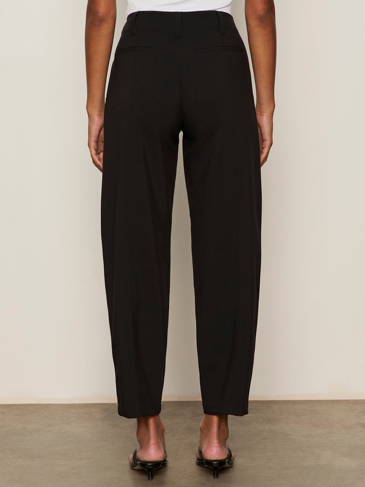 A person stands facing away, wearing Sanctuary Clothing's polished barrel pant in black—high-waisted, relaxed fit, ankle-length—paired with open-toe heels against a plain, neutral background.