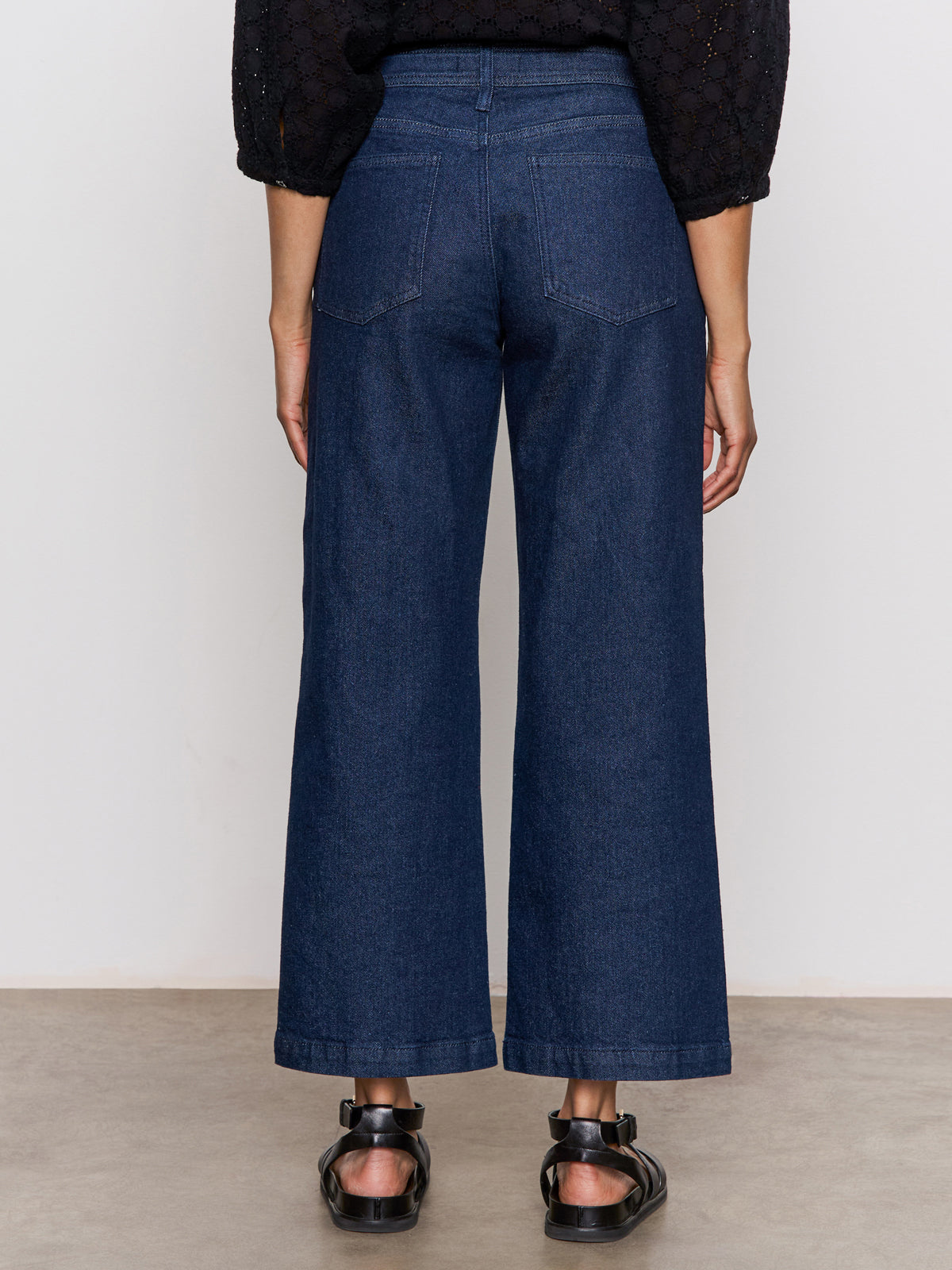 A person stands against a plain background, wearing the Sanctuary Clothing league crop plot twist top with wide-leg blue jeans and black open-toe sandals. The image shows the back view from shoulders to feet.