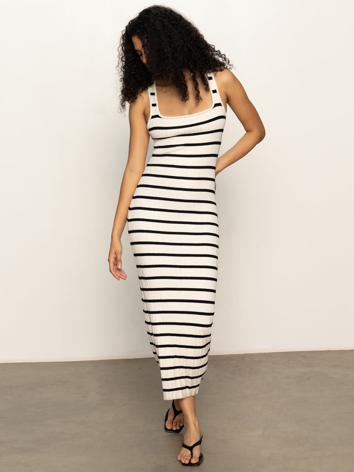 A woman with curly hair wears the Sanctuary Clothing starlit rib maxi light oat black 3, a sleeveless, fitted, black and white striped dress, paired with black sandals. She stands on a concrete floor against a plain white wall.