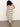 A woman with curly hair wears the Sanctuary Clothing starlit rib maxi light oat black 3, a sleeveless, fitted, black and white striped dress, paired with black sandals. She stands on a concrete floor against a plain white wall.