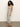 A woman with curly hair stands sideways against a plain background, wearing Sanctuary Clothing's starlit rib maxi light oat black 3, a sleeveless, ankle-length white dress with black stripes and black flip-flops.