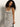 A woman with curly hair models the Sanctuary Clothing starlit rib maxi light oat black 3 dress, featuring a sleeveless, white ribbed design with black stripes, while seated on a stool against an off-white background and smiling slightly at the camera.