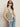 A woman wears Sanctuary Clothing's textured sweater vest macrame black stripe 3 with sunglasses and light blue jeans, posing with one hand in her pocket against a plain light background.