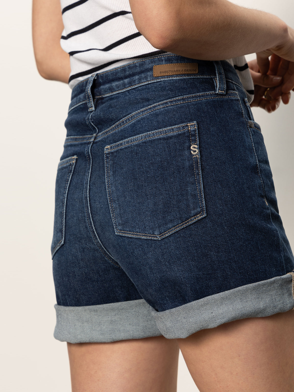 A person in Sanctuary Clothing's cuffed denim short dark with a leather patch and visible back pockets, paired with a striped top, stands in profile to showcase the side and back of their outfit.